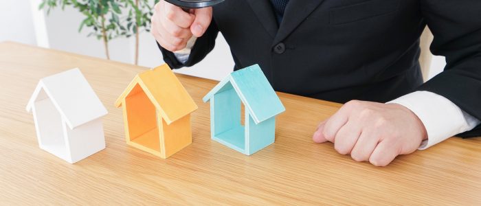 Gutachter hält Lupe über kleine Hausmodelle auf Schreibtisch symbolisch für die Wertermittlung von Immobilien