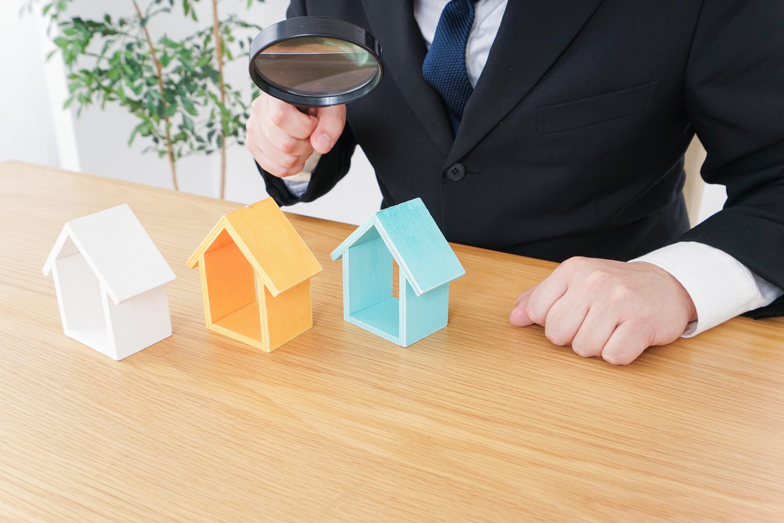 Gutachter hält Lupe über kleine Hausmodelle auf Schreibtisch symbolisch für die Wertermittlung von Immobilien