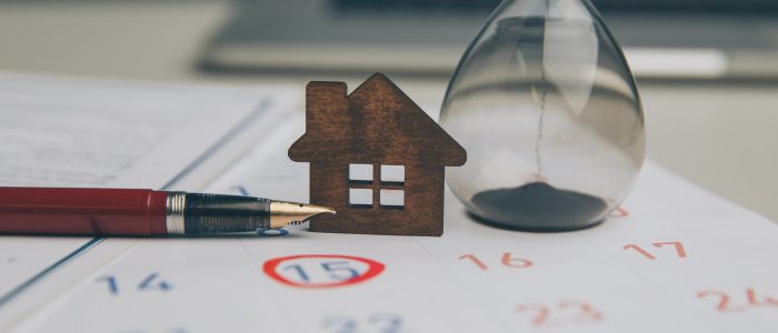 Ein kleines Holzhaus-Modell, ein Füllfederhalter und eine Sanduhr auf einem Kalender mit rot eingekreistem Datum symbolisch für einen Mietkaufvertrag
