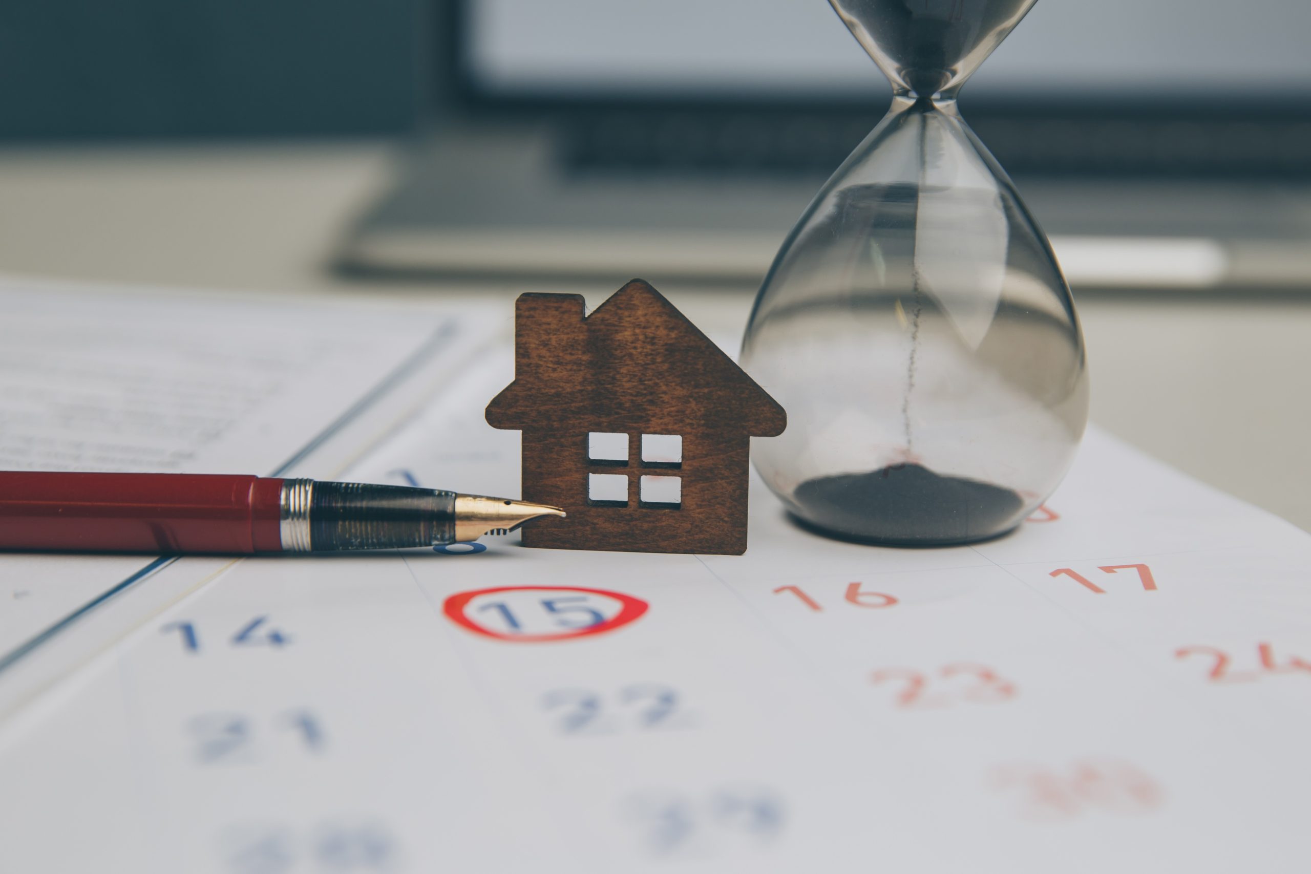 Ein kleines Holzhaus-Modell, ein Füllfederhalter und eine Sanduhr auf einem Kalender mit rot eingekreistem Datum symbolisch für einen Mietkaufvertrag